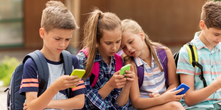 4 Kinder sitzen vor der Schule und schauen auf's Handy. 