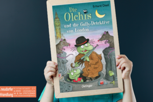 Kind hält eine Tafel mit dem Cover des Buches: Die Olchis und die Gully-Detektive von London