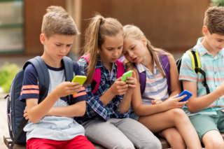 4 Kinder sitzen vor der Schule und schauen auf's Handy. 