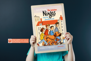 Bub hält eine Tafel mit dem Buchcover con: Die letzten Ninjas und der Juwelenraub; Logo des Sponsors Buchhandlung Neudorfer