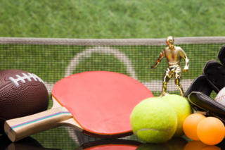Ein Football, ein Tischtennisschläger, Tennisball, Tischtennisbälle und ein Baseball mit Handschuh liegen vor einem grünen Rasenplatz