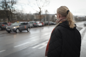 Eine junge Frau steht an einem Straßenrand, man sieht wie ein Taxi zu ihr heranfährt. 