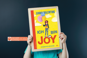 Kind hält eine Tafel hoch mit dem Cover des Buches: Ich bin Joy