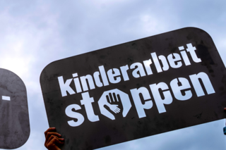 Zwei Hände halten ein Demonstrationsschild mit der Aufschrift "Kinderarbeit stoppen" vor einem blauen Himmel